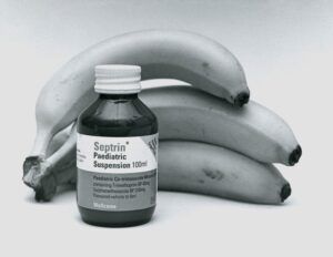 Foto en blanco y negro: un frasco de medicamento pediátrico delante de un manojo de plátanos